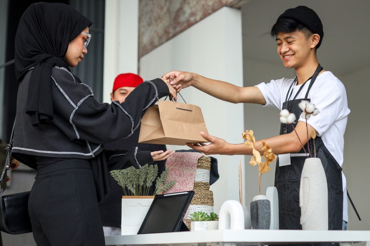 Seorang penjual di kasir sedang melayani pelanggan dengan memberikan produk yang dibeli, mencerminkan peluang usaha modal kecil yang menjanjikan di tahun 2025